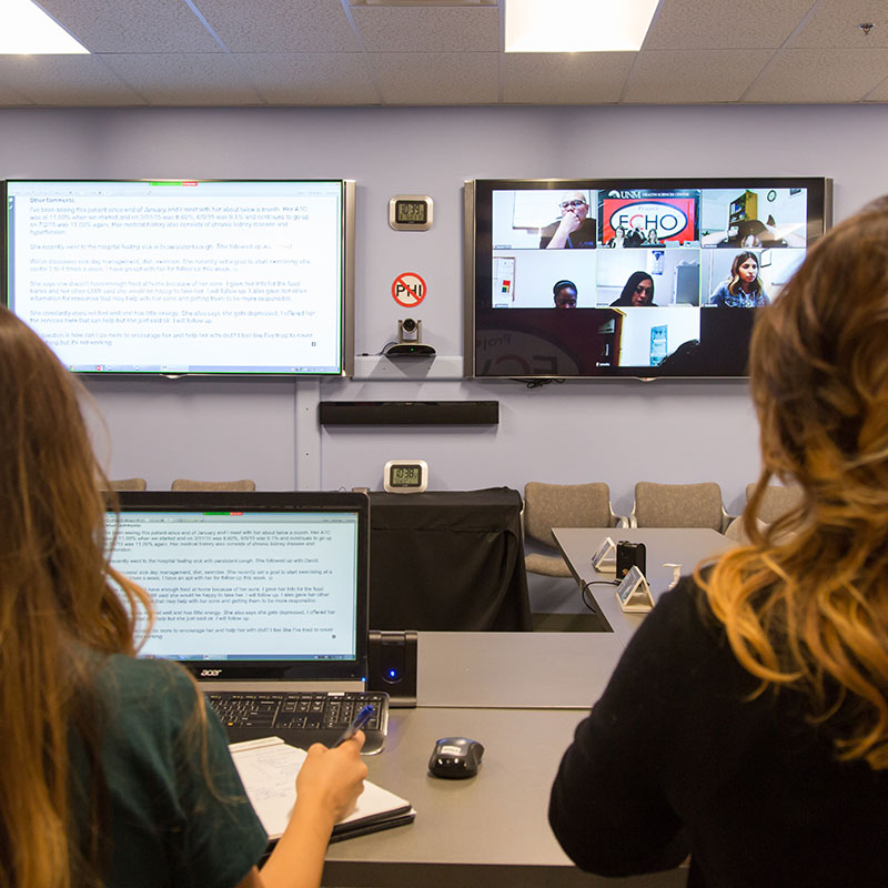 Two women sitting at table in conference room with virtual meeting on TV.